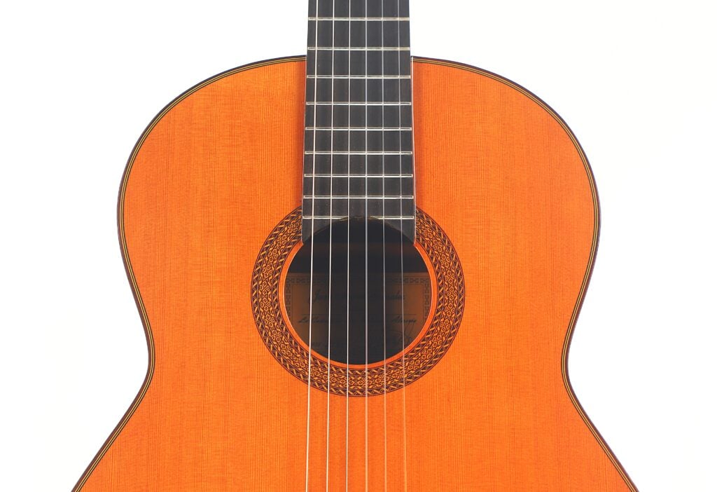 Eine Nahaufnahme von Korpus und Hals der Juan Miguel Gonzalez 2006 Klassische Gitarre mit sechs Saiten und einer dekorativen Rosette um das Schallloch vor weißem Hintergrund. Eine Nahaufnahme von Korpus und Hals der Juan Miguel Gonzalez 2006 Klassische Gitarre mit sechs Saiten und einer dekorativen Rosette um das Schallloch vor weißem Hintergrund.