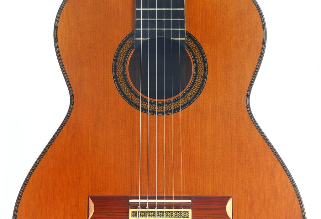Nahaufnahme der „Doble Tapa Klassische Gitarre“ von Manuel Contreras aus dem Jahr 1981, bei der die Saiten, die kunstvoll verzierte Schalllochrosette und der warmbraune Holzkorpus vor weißem Hintergrund besonders hervorgehoben werden. Nahaufnahme der „Doble Tapa Klassische Gitarre“ von Manuel Contreras aus dem Jahr 1981, bei der die Saiten, die kunstvoll verzierte Schalllochrosette und der warmbraune Holzkorpus vor weißem Hintergrund besonders hervorgehoben werden.