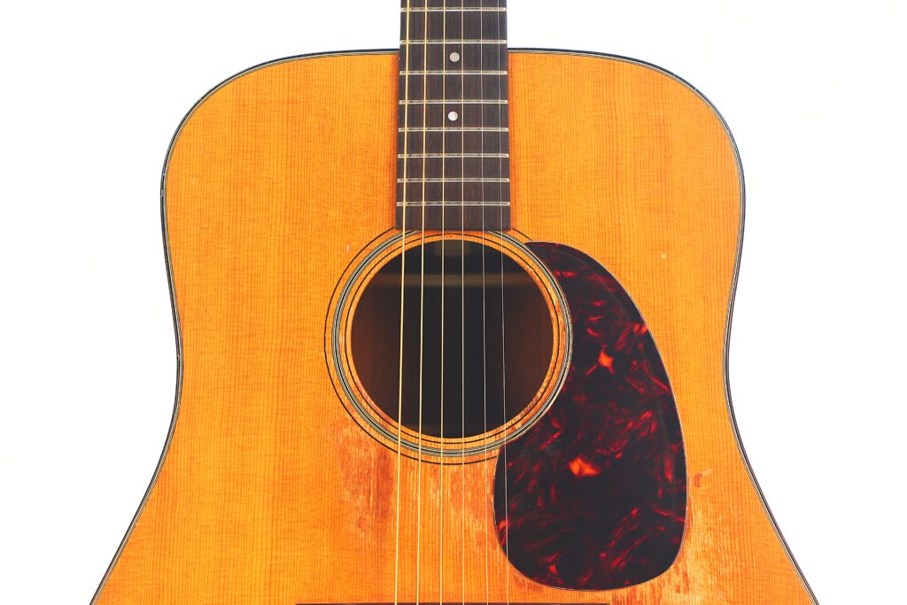 Nahaufnahme einer Martin D-21 1965 Akustik Gitarre, die den hellbraunen Holzkorpus, das schwarz-orangefarbene Schlagbrett, die Stahlsaiten und einen Teil des Halses vor einem schlichten weißen Hintergrund zeigt. Nahaufnahme einer Martin D-21 1965 Akustik Gitarre, die den hellbraunen Holzkorpus, das schwarz-orangefarbene Schlagbrett, die Stahlsaiten und einen Teil des Halses vor einem schlichten weißen Hintergrund zeigt.