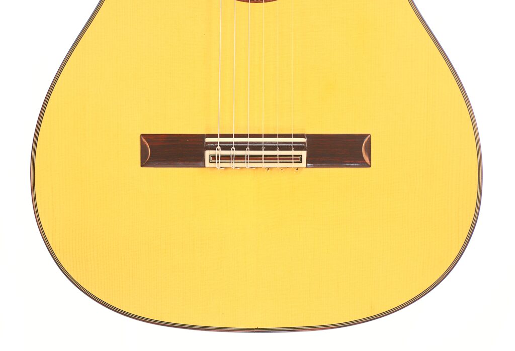 Nahaufnahme der unteren Hälfte einer Vicente Carrillo 1998 Torres Klassische Gitarre. Hervorzuheben sind der helle Holzkorpus, die Saiten und der dunkle Holzsteg. Nahaufnahme der unteren Hälfte einer Vicente Carrillo 1998 Torres Klassische Gitarre. Hervorzuheben sind der helle Holzkorpus, die Saiten und der dunkle Holzsteg.