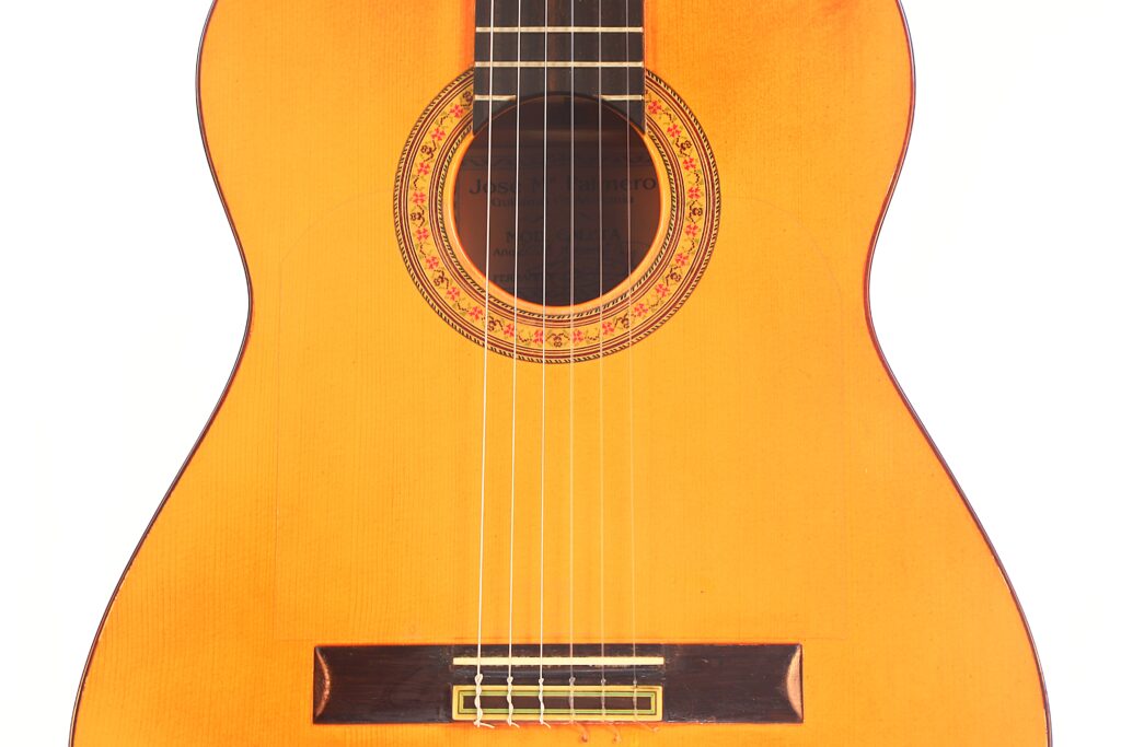 Eine Nahaufnahme der Flamenco-Gitarre „José M Palmero 2023“ zeigt das Schallloch mit einer dekorativen Rosette, sechs Saiten und einen Steg aus Holz, alles vor einem weißen Hintergrund. Eine Nahaufnahme der Flamenco-Gitarre „José M Palmero 2023“ zeigt das Schallloch mit einer dekorativen Rosette, sechs Saiten und einen Steg aus Holz, alles vor einem weißen Hintergrund.