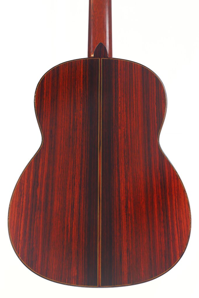 Das Bild zeigt die Rückseite der „Klassische Gitarre“ von Joachim Schneider aus dem Jahr 1977, die aus poliertem dunklem Holz mit rotbraunen Streifen, einer Mittelnaht und einer glänzenden Oberfläche gefertigt ist. Das Bild zeigt die Rückseite der „Klassische Gitarre“ von Joachim Schneider aus dem Jahr 1977, die aus poliertem dunklem Holz mit rotbraunen Streifen, einer Mittelnaht und einer glänzenden Oberfläche gefertigt ist.