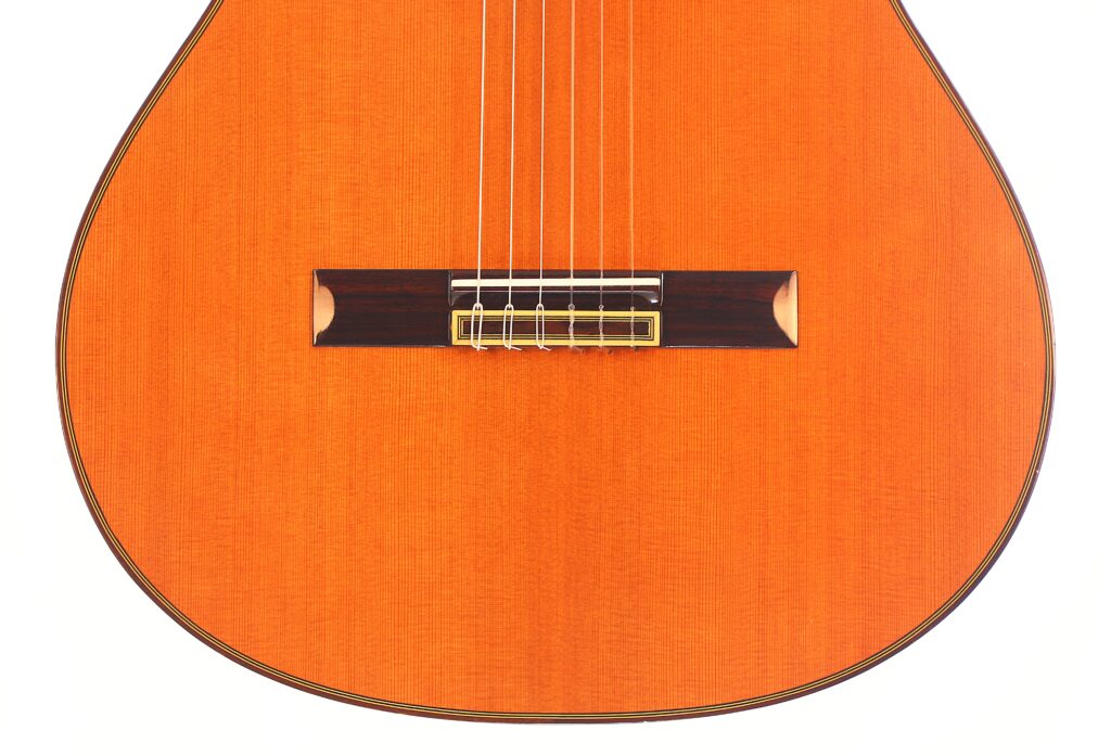 Nahaufnahme der unteren Hälfte einer Juan Miguel Gonzalez 2006 Klassische Gitarre, wobei der Holzkorpus, der Steg und die sechs Saiten vor einem weißen Hintergrund hervorgehoben werden. Nahaufnahme der unteren Hälfte einer Juan Miguel Gonzalez 2006 Klassische Gitarre, wobei der Holzkorpus, der Steg und die sechs Saiten vor einem weißen Hintergrund hervorgehoben werden.