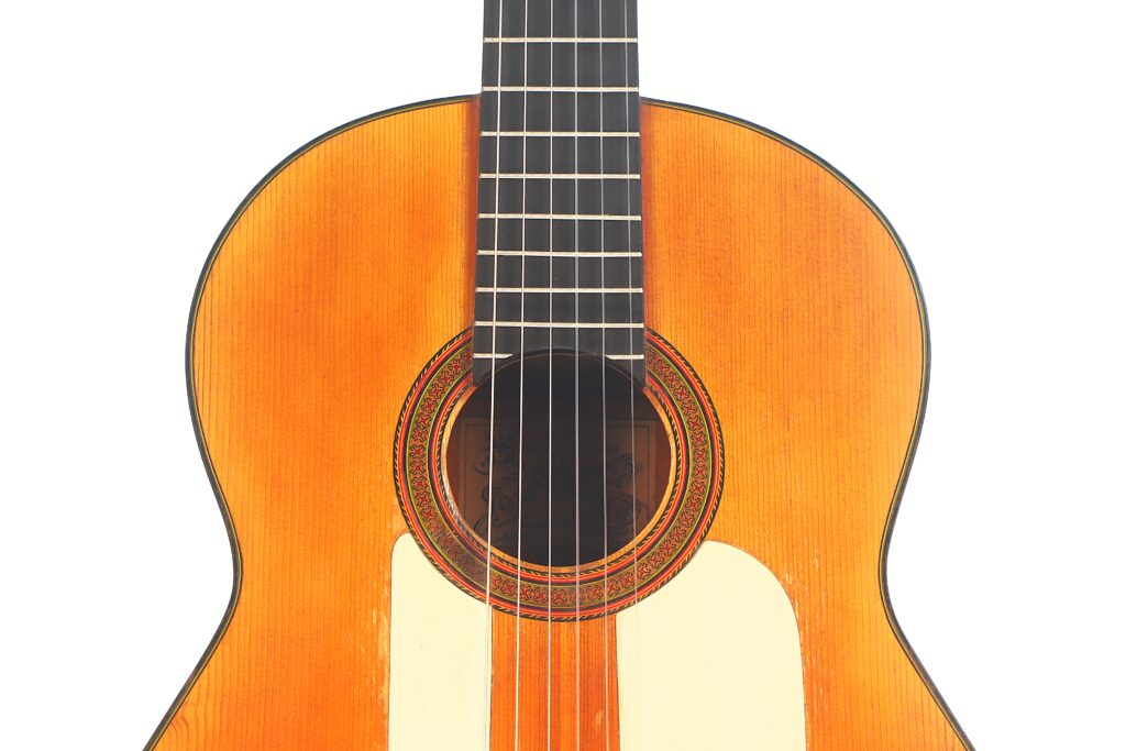 Nahaufnahme einer Flamenco-Gitarre von Jose Ramirez aus dem Jahr 1955. Hervorzuheben sind die sechs Saiten, der hölzerne Resonanzboden mit einer dekorativen Rosette, die glänzende Oberfläche und der schlichte weiße Hintergrund. Nahaufnahme einer Flamenco-Gitarre von Jose Ramirez aus dem Jahr 1955. Hervorzuheben sind die sechs Saiten, der hölzerne Resonanzboden mit einer dekorativen Rosette, die glänzende Oberfläche und der schlichte weiße Hintergrund.