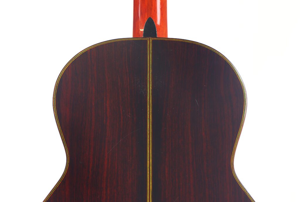 Das Bild zeigt die Rückseite einer Juan Estruch Chet Atkins Yellow Label 1976 Klassische Gitarre mit dunkler Holzoberfläche, einem zentralen Einlegeband, heller Einfassung und einem rötlichen Hals, der sich vom Korpus abhebt, vor einem schlichten weißen Hintergrund. Das Bild zeigt die Rückseite einer Juan Estruch Chet Atkins Yellow Label 1976 Klassische Gitarre mit dunkler Holzoberfläche, einem zentralen Einlegeband, heller Einfassung und einem rötlichen Hals, der sich vom Korpus abhebt, vor einem schlichten weißen Hintergrund.