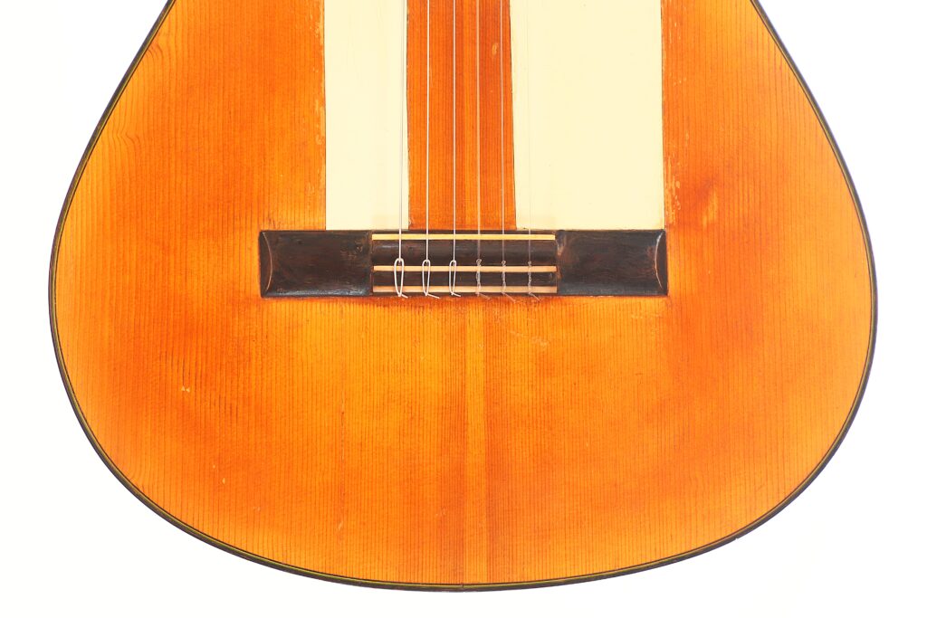 Nahaufnahme des Korpus einer Flamenco-Gitarre von Jose Ramirez aus dem Jahr 1955. Hervorzuheben sind die Saiten, der Steg, die warme orange-braune Holzmaserung und der Mittelstreifen aus hellerem Holz. Nahaufnahme des Korpus einer Flamenco-Gitarre von Jose Ramirez aus dem Jahr 1955. Hervorzuheben sind die Saiten, der Steg, die warme orange-braune Holzmaserung und der Mittelstreifen aus hellerem Holz.
