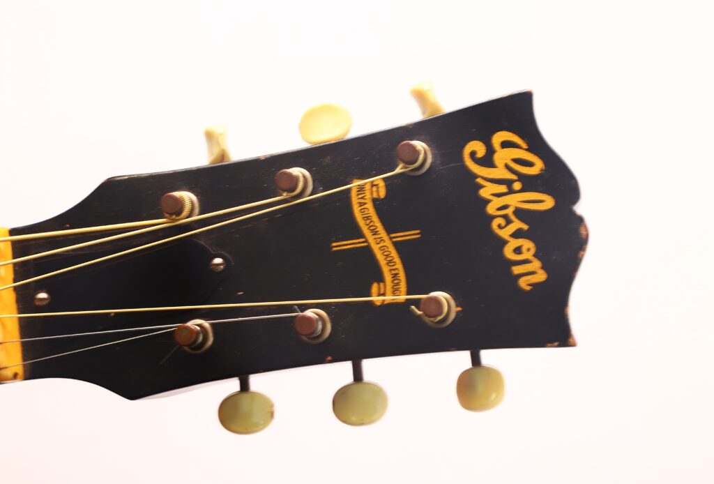 Nahaufnahme der Kopfplatte einer schwarzen Gibson J-45 Banner 1944 Akustikgitarre mit sichtbaren Stimmwirbeln und Saiten vor hellem Hintergrund. Das gelbe Gibson-Logo hebt sich deutlich ab. Nahaufnahme der Kopfplatte einer schwarzen Gibson J-45 Banner 1944 Akustikgitarre mit sichtbaren Stimmwirbeln und Saiten vor hellem Hintergrund. Das gelbe Gibson-Logo hebt sich deutlich ab.