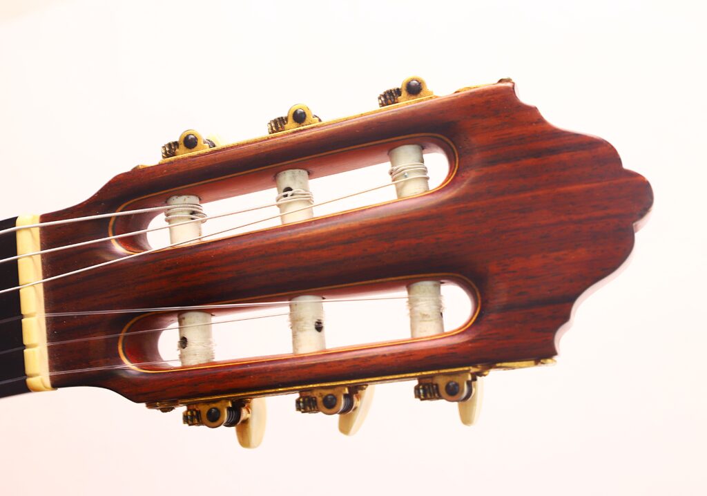 Nahaufnahme der Kopfplatte der Juan Montero 1993 Blanca Flamenco Gitarre mit hölzernen Stimmwirbeln, offenen Schlitzen und Metallstimmmechaniken vor einem schlichten hellen Hintergrund. Nahaufnahme der Kopfplatte der Juan Montero 1993 Blanca Flamenco Gitarre mit hölzernen Stimmwirbeln, offenen Schlitzen und Metallstimmmechaniken vor einem schlichten hellen Hintergrund.