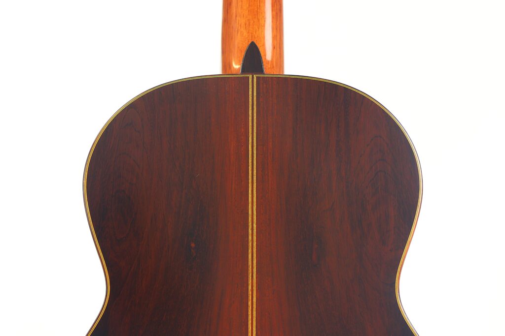Nahaufnahme der Rückseite der Antonio Aparicio Mod. Master Klassische Gitarre, mit dunklem Holz mit sichtbarer Maserung und einer dünnen vertikalen Ziereinlage. Der Hals der Gitarre ist an der Decke teilweise sichtbar. Nahaufnahme der Rückseite der Antonio Aparicio Mod. Master Klassische Gitarre, mit dunklem Holz mit sichtbarer Maserung und einer dünnen vertikalen Ziereinlage. Der Hals der Gitarre ist an der Decke teilweise sichtbar.