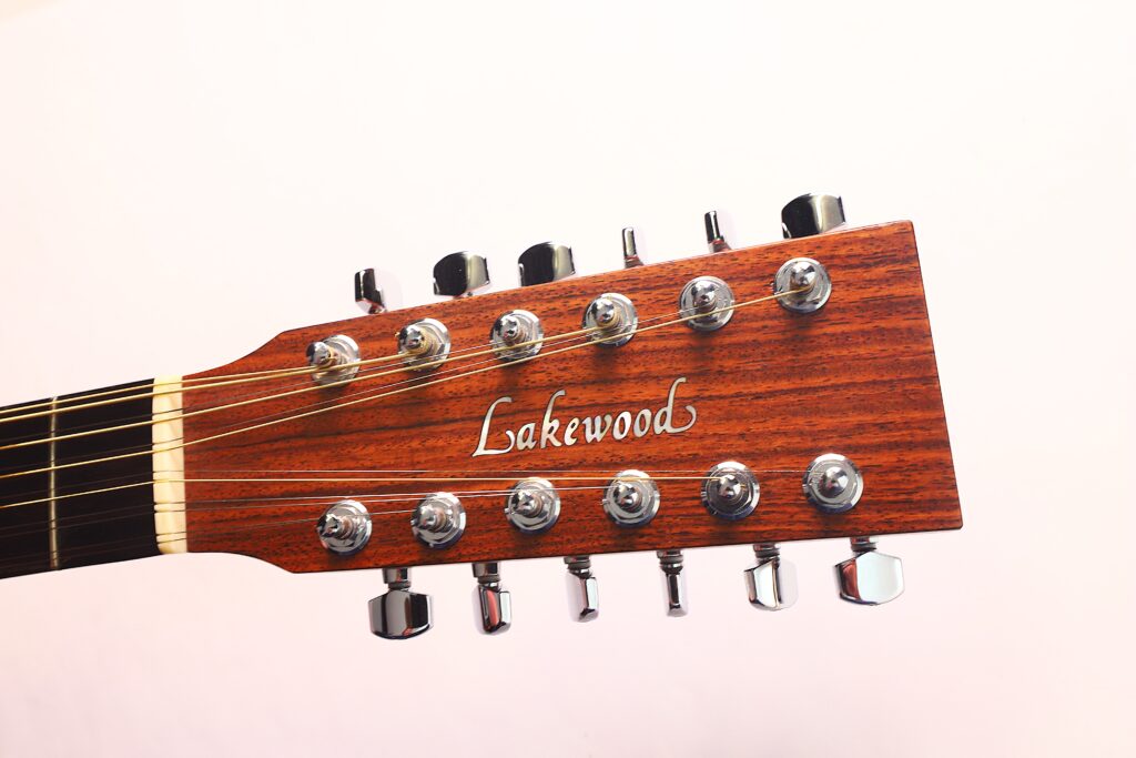 Nahaufnahme der Kopfplatte einer Lakewood D-14-12 Cut 2005 Akustik-Gitarre, mit Stimmwirbeln, Saiten und dem Lakewood-Logo auf Holz vor hellem Hintergrund. Nahaufnahme der Kopfplatte einer Lakewood D-14-12 Cut 2005 Akustik-Gitarre, mit Stimmwirbeln, Saiten und dem Lakewood-Logo auf Holz vor hellem Hintergrund.