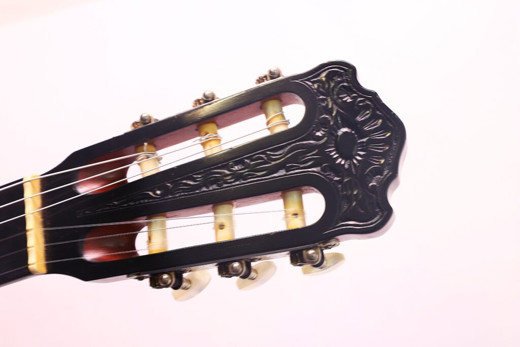 Nahaufnahme des Kopfes der „Joachim Schneider 1977 Klassische Gitarre“ mit kunstvollen schwarzen Verzierungen, weißen Stimmwirbeln und Nylonsaiten vor einem hellrosa Hintergrund. Nahaufnahme des Kopfes der „Joachim Schneider 1977 Klassische Gitarre“ mit kunstvollen schwarzen Verzierungen, weißen Stimmwirbeln und Nylonsaiten vor einem hellrosa Hintergrund.
