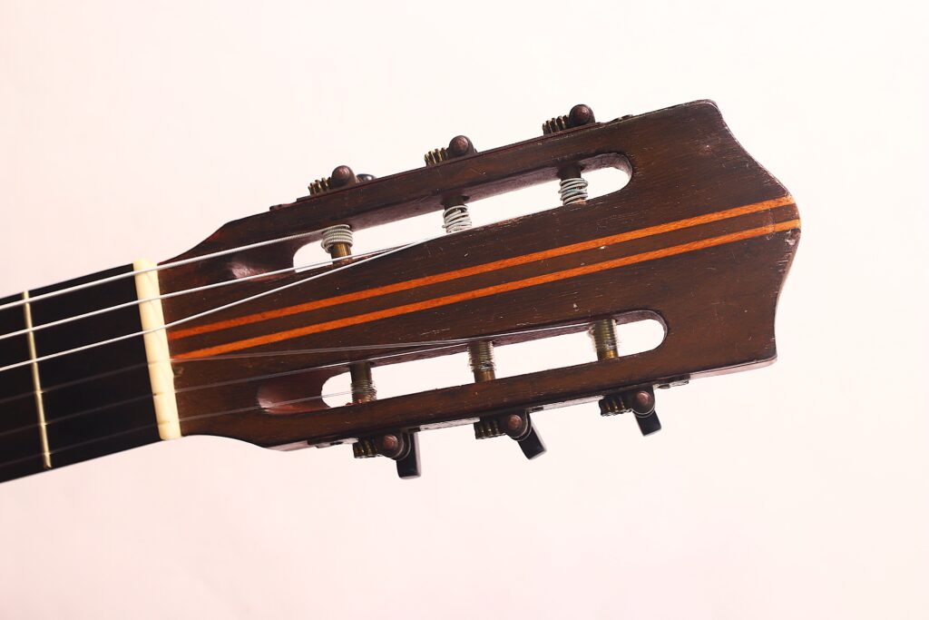 Nahaufnahme der Kopfplatte der Andres Marin 1900 Klassische Gitarre mit sechs Stimmwirbeln (drei pro Seite) und zwei orangefarbenen Streifen auf dem Holz. Der Hintergrund ist einfarbig und hell gefärbt. Nahaufnahme der Kopfplatte der Andres Marin 1900 Klassische Gitarre mit sechs Stimmwirbeln (drei pro Seite) und zwei orangefarbenen Streifen auf dem Holz. Der Hintergrund ist einfarbig und hell gefärbt.