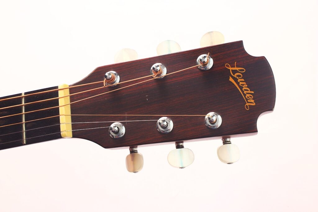 Nahaufnahme der Kopfplatte der Lowden S-5 Akustik Gitarre mit sechs Metallstimmwirbeln, elfenbeinfarbenen Stimmknöpfen und dem goldenen Lowden-Logo auf hellem Grund. Nahaufnahme der Kopfplatte der Lowden S-5 Akustik Gitarre mit sechs Metallstimmwirbeln, elfenbeinfarbenen Stimmknöpfen und dem goldenen Lowden-Logo auf hellem Grund.