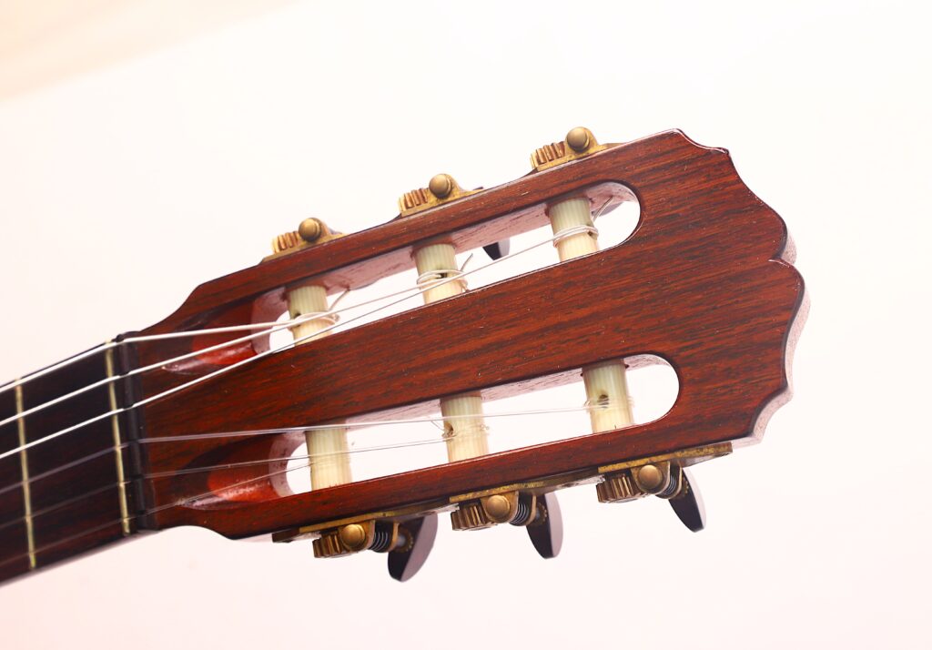 Nahaufnahme des Kopfes der „Carl Hermann Schäfer – Nicolaus Wollf 2012 Klassische Gitarre“, die die Stimmwirbel, Saiten und die Holzoberfläche vor einem schlichten, hellen Hintergrund zeigt. Nahaufnahme des Kopfes der „Carl Hermann Schäfer – Nicolaus Wollf 2012 Klassische Gitarre“, die die Stimmwirbel, Saiten und die Holzoberfläche vor einem schlichten, hellen Hintergrund zeigt.