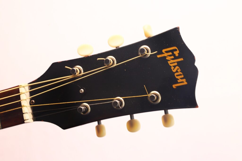 Nahaufnahme des Kopfes der Gibson J-50 Akustikgitarre aus dem Jahr 1952 mit Stimmwirbeln und gelbem Logo vor einem schlichten, hellen Hintergrund. Nahaufnahme des Kopfes der Gibson J-50 Akustikgitarre aus dem Jahr 1952 mit Stimmwirbeln und gelbem Logo vor einem schlichten, hellen Hintergrund.