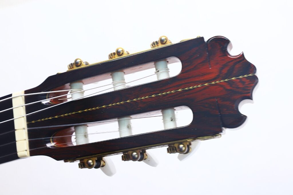Nahaufnahme des Kopfes einer „Doble Tapa Klassische Gitarre“ von Manuel Contreras aus dem Jahr 1981, mit Stimmwirbeln, weißen Saiten und einer dunklen Holzoberfläche mit aufwendigen Details vor einem schlichten weißen Hintergrund. Nahaufnahme des Kopfes einer „Doble Tapa Klassische Gitarre“ von Manuel Contreras aus dem Jahr 1981, mit Stimmwirbeln, weißen Saiten und einer dunklen Holzoberfläche mit aufwendigen Details vor einem schlichten weißen Hintergrund.