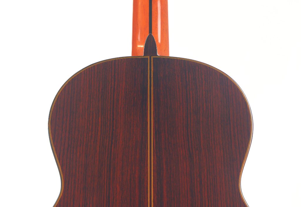 Nahaufnahme der Rückseite einer Juan Miguel Gonzalez 2006 Klassische Gitarre, die eine dunkle Holzmaserung mit einer zentralen Naht und einem hellen Einband vor weißem Hintergrund zeigt. Nahaufnahme der Rückseite einer Juan Miguel Gonzalez 2006 Klassische Gitarre, die eine dunkle Holzmaserung mit einer zentralen Naht und einem hellen Einband vor weißem Hintergrund zeigt.