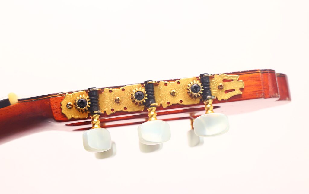 Nahaufnahme der Stimmwirbel und Zahnräder am Kopf einer Juan Estruch Chet Atkins Yellow Label 1976 Klassische Gitarre, die drei weiße Stimmknöpfe und verzierte Goldplatten vor einem schlichten weißen Hintergrund zeigt. Nahaufnahme der Stimmwirbel und Zahnräder am Kopf einer Juan Estruch Chet Atkins Yellow Label 1976 Klassische Gitarre, die drei weiße Stimmknöpfe und verzierte Goldplatten vor einem schlichten weißen Hintergrund zeigt.