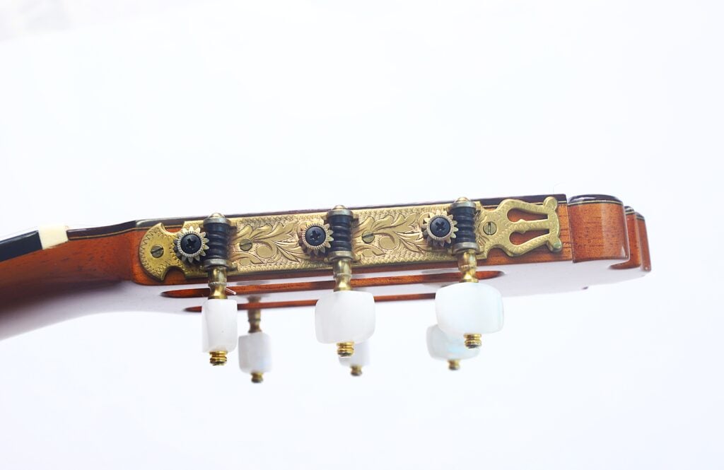 Nahaufnahme des Kopfes einer „Doble Tapa Klassische Gitarre“ von Manuel Contreras aus dem Jahr 1981 mit kunstvoll verzierten goldenen Stimmwirbeln und weißen Stimmknöpfen vor einem schlichten weißen Hintergrund. Nahaufnahme des Kopfes einer „Doble Tapa Klassische Gitarre“ von Manuel Contreras aus dem Jahr 1981 mit kunstvoll verzierten goldenen Stimmwirbeln und weißen Stimmknöpfen vor einem schlichten weißen Hintergrund.