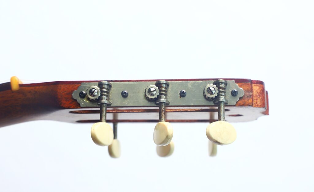 Nahaufnahme der Stimmwirbel am Kopf einer Jose Ramirez I 1921 Klassische Gitarre, mit einer Metallplatte und drei cremefarbenen Stimmwirbeln vor einem schlichten weißen Hintergrund. Nahaufnahme der Stimmwirbel am Kopf einer Jose Ramirez I 1921 Klassische Gitarre, mit einer Metallplatte und drei cremefarbenen Stimmwirbeln vor einem schlichten weißen Hintergrund.