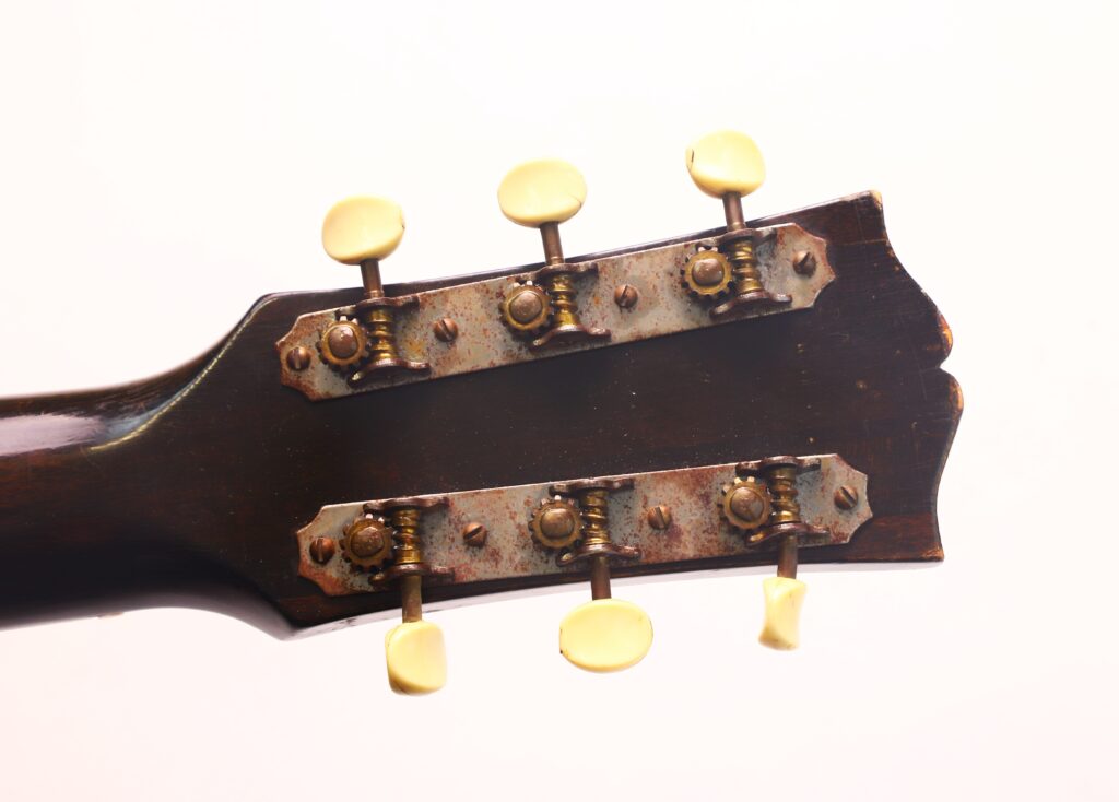 Nahaufnahme der Kopfplatte einer Gibson J-45 Banner Akustikgitarre aus dem Jahr 1944 mit sechs gelben Stimmwirbeln und Metallmechanismen vor einem schlichten weißen Hintergrund. Nahaufnahme der Kopfplatte einer Gibson J-45 Banner Akustikgitarre aus dem Jahr 1944 mit sechs gelben Stimmwirbeln und Metallmechanismen vor einem schlichten weißen Hintergrund.