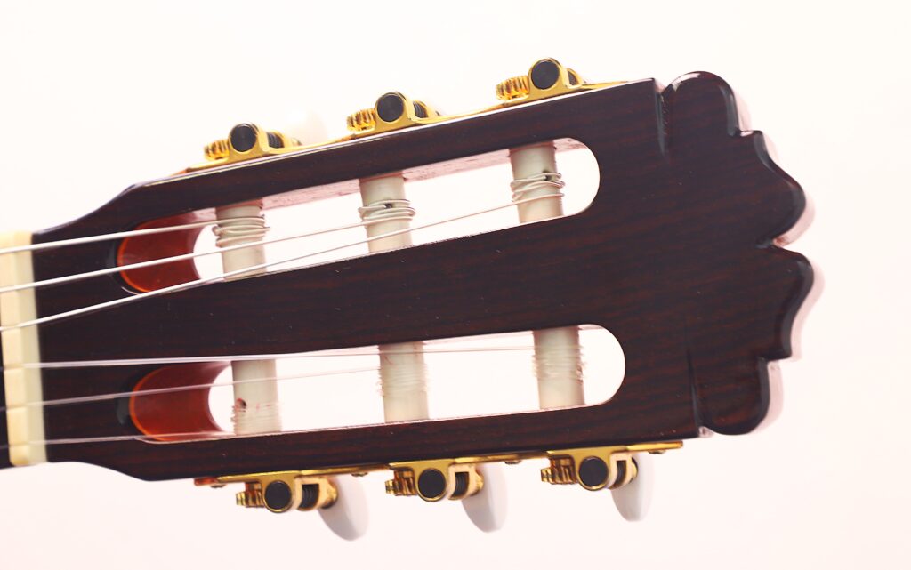 Nahaufnahme der Kopfplatte der Juan Miguel Gonzalez 2006 Klassische Gitarre mit Stimmwirbeln, Saiten und einer dunklen Holzoberfläche mit goldenen Beschlägen vor einem hellen Hintergrund. Nahaufnahme der Kopfplatte der Juan Miguel Gonzalez 2006 Klassische Gitarre mit Stimmwirbeln, Saiten und einer dunklen Holzoberfläche mit goldenen Beschlägen vor einem hellen Hintergrund.