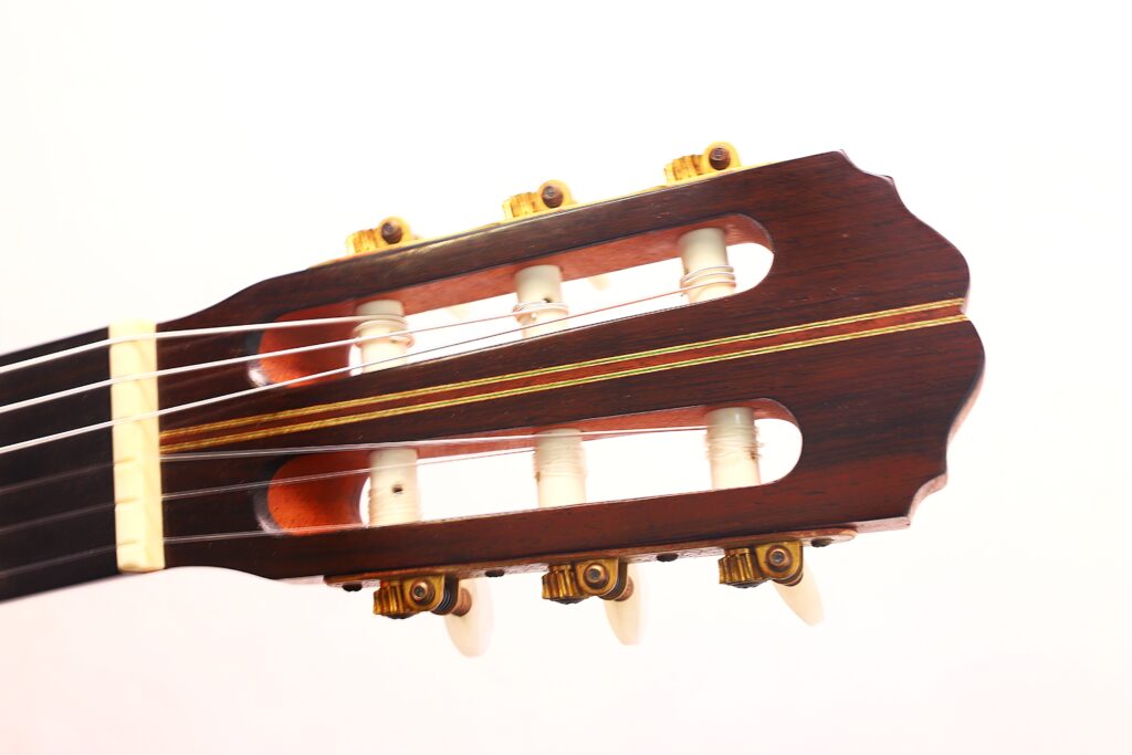 Nahaufnahme der Kopfplatte der Antonio Aparicio Mod. Master Klassische Gitarre mit sechs Stimmwirbeln, Nylonsaiten und einer Holzoberfläche vor einem schlichten weißen Hintergrund. Nahaufnahme der Kopfplatte der Antonio Aparicio Mod. Master Klassische Gitarre mit sechs Stimmwirbeln, Nylonsaiten und einer Holzoberfläche vor einem schlichten weißen Hintergrund.