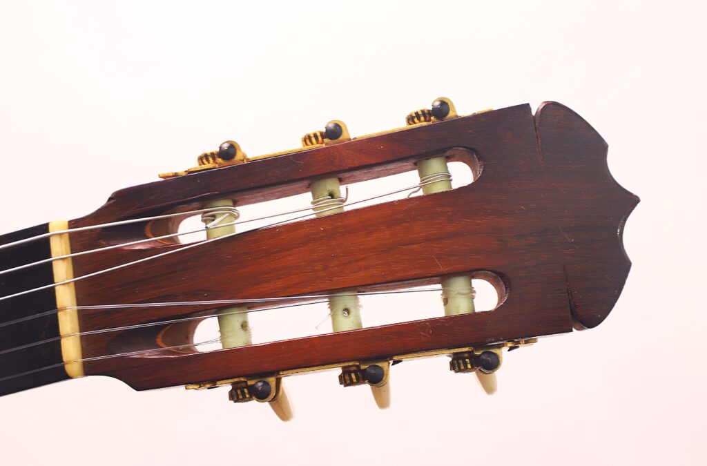 Nahaufnahme der Kopfplatte einer Flamenco-Gitarre von Jose Ramirez aus dem Jahr 1955, bei der Stimmwirbel, Saiten und Holzmaserung vor einem hellrosa Hintergrund sichtbar sind. Nahaufnahme der Kopfplatte einer Flamenco-Gitarre von Jose Ramirez aus dem Jahr 1955, bei der Stimmwirbel, Saiten und Holzmaserung vor einem hellrosa Hintergrund sichtbar sind.