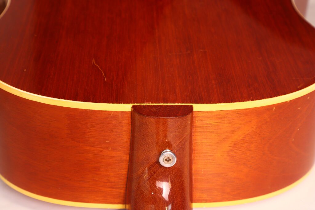 Nahaufnahme des Rückens und der Zargen der Gibson J-50 Akustikgitarre aus dem Jahr 1952, die die glänzende Holzoberfläche, die gelbe Einfassung, den Halsansatz und den angebrachten Gurtknopf hervorhebt. Nahaufnahme des Rückens und der Zargen der Gibson J-50 Akustikgitarre aus dem Jahr 1952, die die glänzende Holzoberfläche, die gelbe Einfassung, den Halsansatz und den angebrachten Gurtknopf hervorhebt.