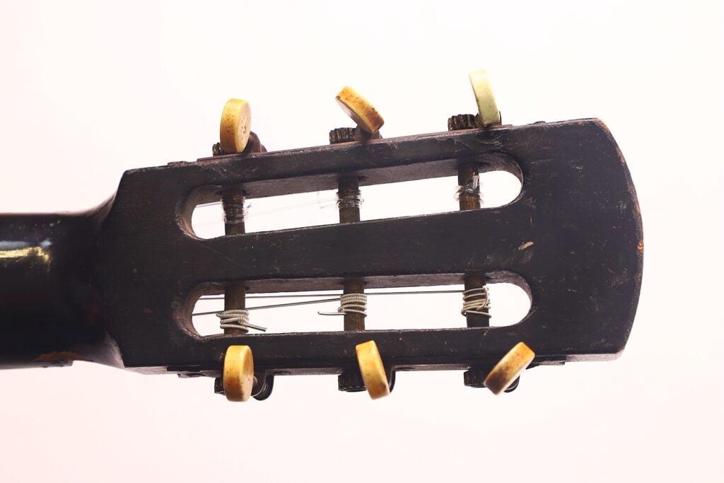 Nahaufnahme der Kopfplatte der Klassischen Gitarre von Braun & Hauser aus dem Jahr 1905 mit sechs Stimmwirbeln und Saiten vor einem schlichten hellen Hintergrund. Das abgenutzte Holz deutet auf das Alter und den häufigen Gebrauch hin. Nahaufnahme der Kopfplatte der Klassischen Gitarre von Braun & Hauser aus dem Jahr 1905 mit sechs Stimmwirbeln und Saiten vor einem schlichten hellen Hintergrund. Das abgenutzte Holz deutet auf das Alter und den häufigen Gebrauch hin.