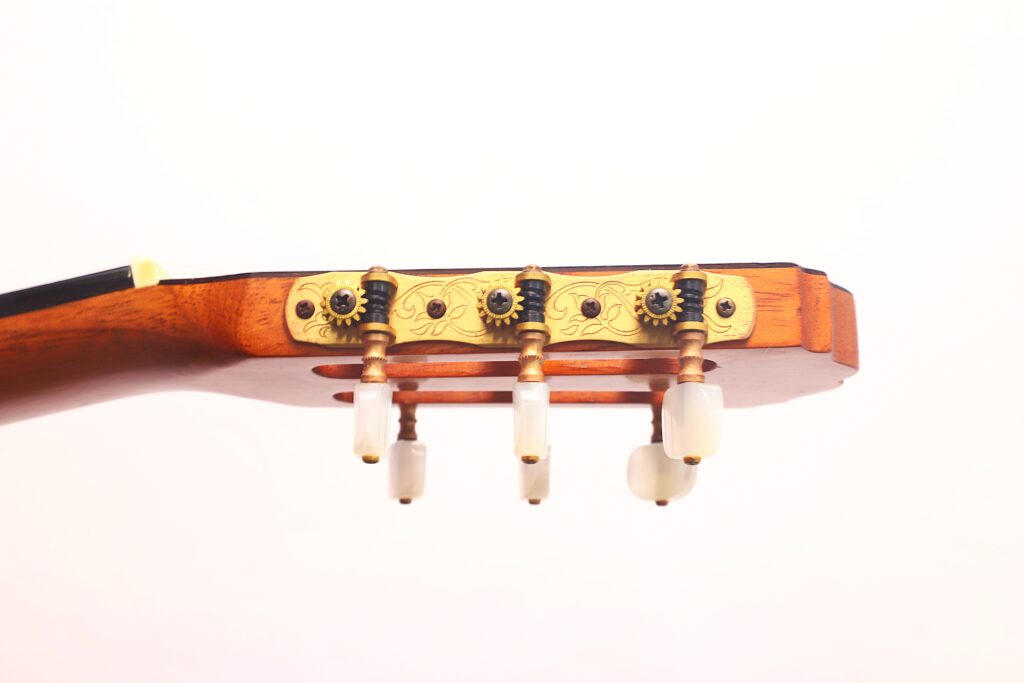Nahaufnahme der Stimmwirbel und der Kopfplatte der Antonio Aparicio Mod. Master Klassische Gitarre, mit goldfarbenen verzierten Metallplatten und weißen Stimmknöpfen vor einem schlichten weißen Hintergrund. Nahaufnahme der Stimmwirbel und der Kopfplatte der Antonio Aparicio Mod. Master Klassische Gitarre, mit goldfarbenen verzierten Metallplatten und weißen Stimmknöpfen vor einem schlichten weißen Hintergrund.
