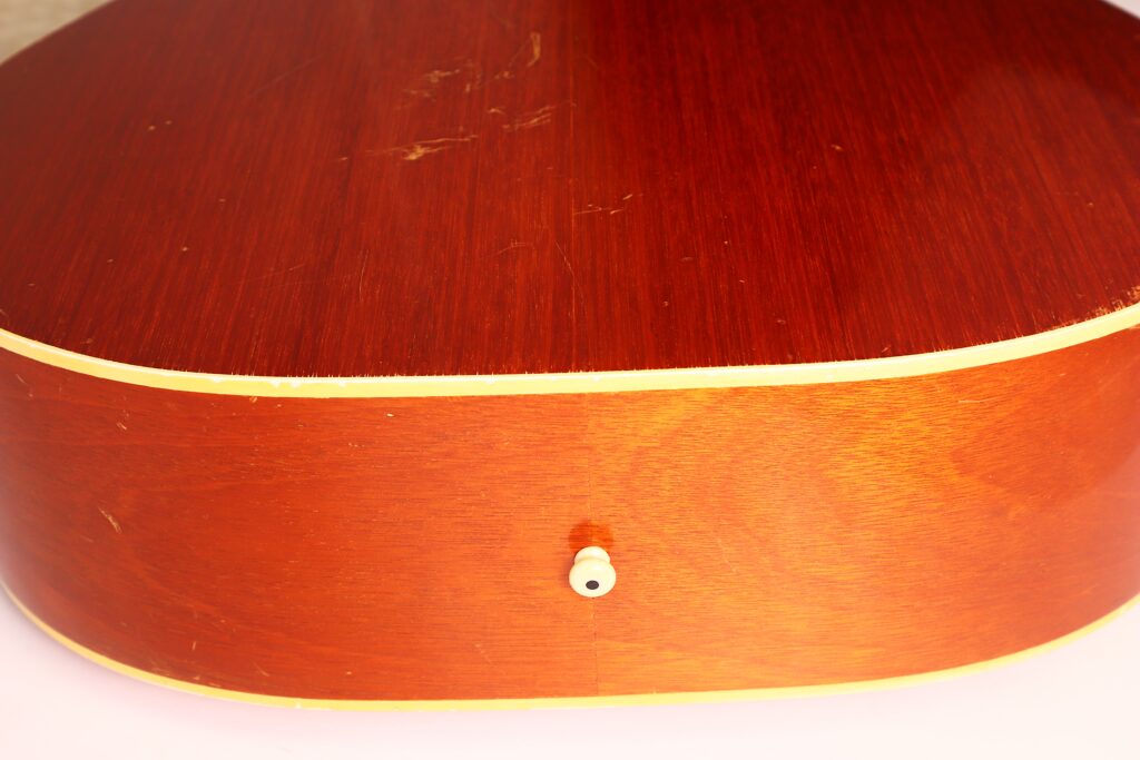 Nahaufnahme der Unterkante der Gibson J-50 Akustikgitarre aus dem Jahr 1952, die die Holzmaserung, die cremefarbene Einfassung und den weißen Gurtknopf unten in der Mitte zeigt. Nahaufnahme der Unterkante der Gibson J-50 Akustikgitarre aus dem Jahr 1952, die die Holzmaserung, die cremefarbene Einfassung und den weißen Gurtknopf unten in der Mitte zeigt.