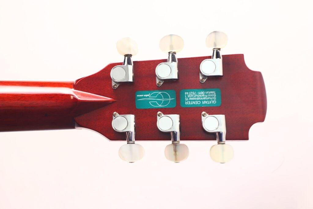 Nahaufnahme der Rückseite einer Lowden S-5 Akustik Gitarre mit sechs Stimmwirbeln, rötlicher Holzoberfläche und einem grünen Guitar Center-Aufkleber auf hellem Grund. Nahaufnahme der Rückseite einer Lowden S-5 Akustik Gitarre mit sechs Stimmwirbeln, rötlicher Holzoberfläche und einem grünen Guitar Center-Aufkleber auf hellem Grund.