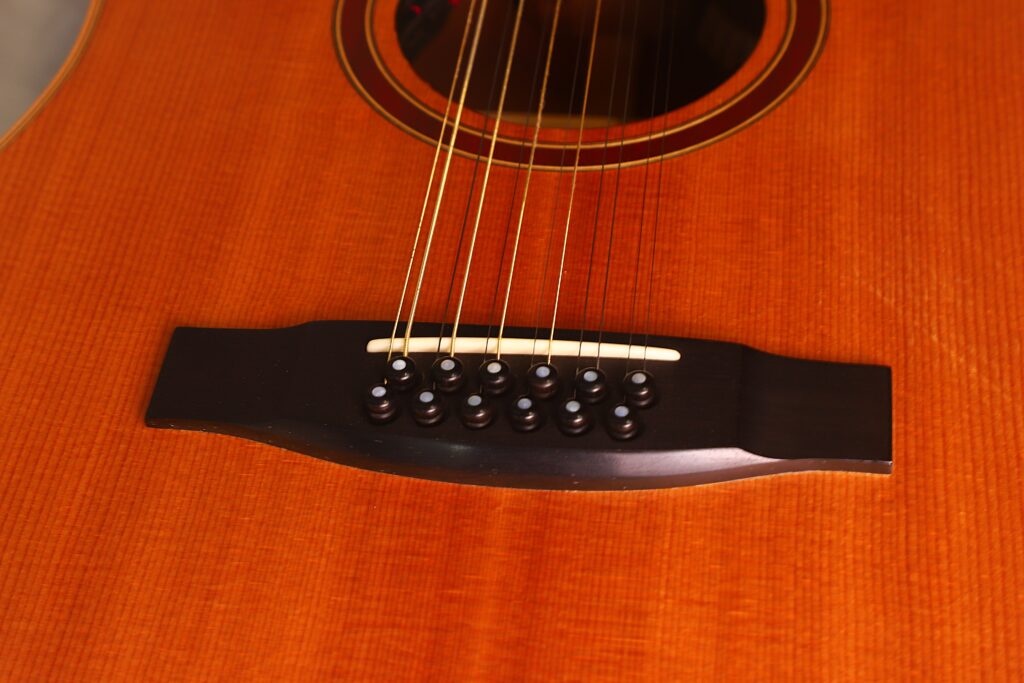 Nahaufnahme des Stegs und der Saiten der Lakewood D-14-12 Cut 2005 Akustik Gitarre, die die Holzmaserung und die detaillierte Handwerkskunst zeigt. Nahaufnahme des Stegs und der Saiten der Lakewood D-14-12 Cut 2005 Akustik Gitarre, die die Holzmaserung und die detaillierte Handwerkskunst zeigt.