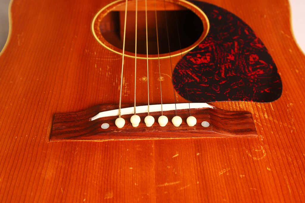 Nahaufnahme des unteren Teils der Gibson J-50 Akustikgitarre aus dem Jahr 1952, die den Holzsteg, die sechs Saiten mit weißen Stegstiften und ein rot-schwarzes Schlagbrett auf einem abgenutzten braunen Lack hervorhebt. Nahaufnahme des unteren Teils der Gibson J-50 Akustikgitarre aus dem Jahr 1952, die den Holzsteg, die sechs Saiten mit weißen Stegstiften und ein rot-schwarzes Schlagbrett auf einem abgenutzten braunen Lack hervorhebt.