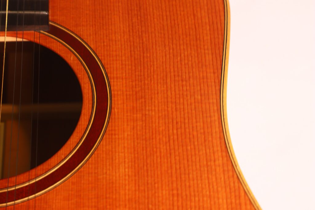 Nahaufnahme des Korpus einer Lakewood D-14-12 Cut 2005 Akustik Gitarre. Hervorzuheben sind das Schallloch, die Holzmaserung und die Saiten auf der linken Seite vor einem weißen Hintergrund auf der rechten Seite. Der warme Ton der Gitarre hebt sich im Kontrast ab. Nahaufnahme des Korpus einer Lakewood D-14-12 Cut 2005 Akustik Gitarre. Hervorzuheben sind das Schallloch, die Holzmaserung und die Saiten auf der linken Seite vor einem weißen Hintergrund auf der rechten Seite. Der warme Ton der Gitarre hebt sich im Kontrast ab.