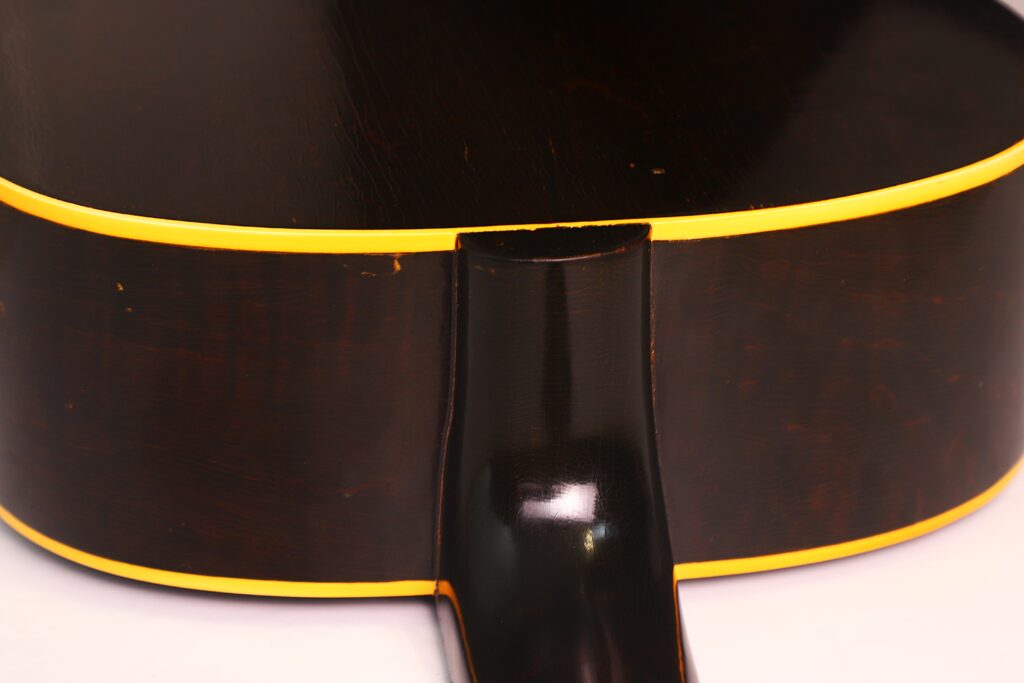 Nahaufnahme des Bodens und der Halsverbindung der Gibson J-45 Banner 1944 Akustik Gitarre. Hervorzuheben sind die dunkelbraune Lackierung mit gelber Zierleiste, die glatte, glänzende Oberfläche und der nahtlose Übergang zwischen Hals und Korpus. Nahaufnahme des Bodens und der Halsverbindung der Gibson J-45 Banner 1944 Akustik Gitarre. Hervorzuheben sind die dunkelbraune Lackierung mit gelber Zierleiste, die glatte, glänzende Oberfläche und der nahtlose Übergang zwischen Hals und Korpus.