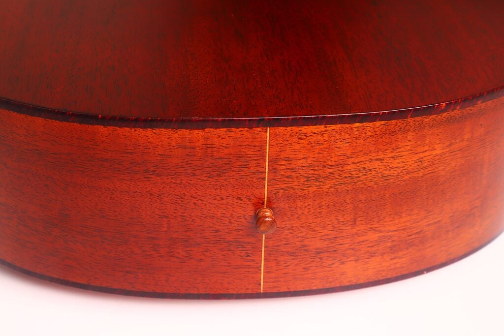 Nahaufnahme der Rückseite und der Seite einer Lowden S-5 Akustikgitarre mit polierter rötlich-brauner Holzmaserung, einem kleinen Endstift und einer dünnen hellen vertikalen Naht. Nahaufnahme der Rückseite und der Seite einer Lowden S-5 Akustikgitarre mit polierter rötlich-brauner Holzmaserung, einem kleinen Endstift und einer dünnen hellen vertikalen Naht.