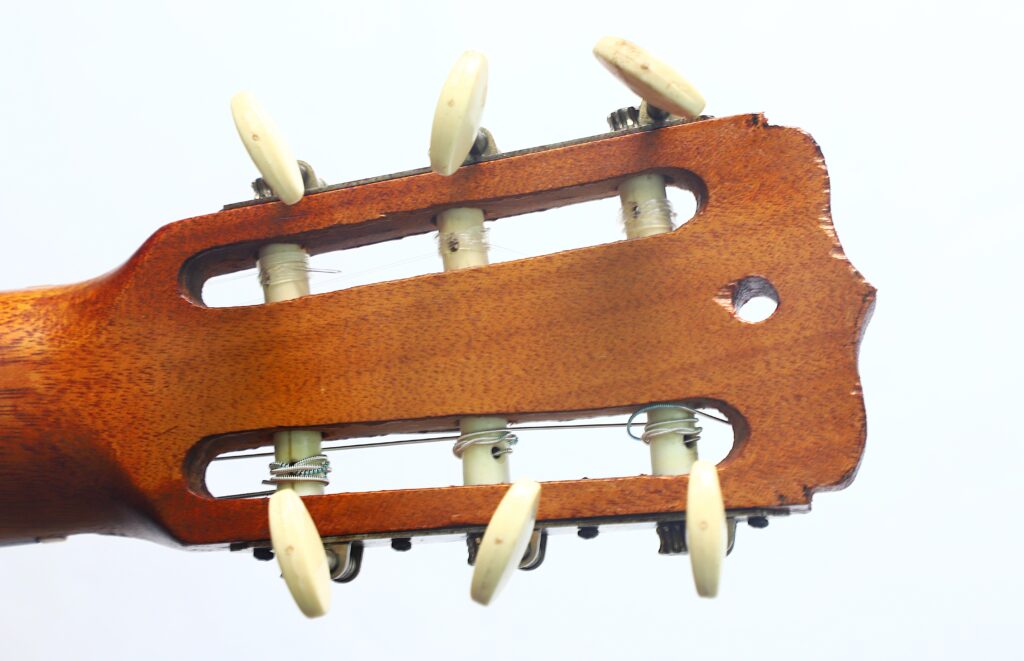 Nahaufnahme des Kopfstücks einer Jose Ramirez I 1921 Klassische Gitarre, die die abgenutzten Stimmwirbel und die abgeplatzten Stellen am Rand hervorhebt, vor einem schlichten weißen Hintergrund. Nahaufnahme des Kopfstücks einer Jose Ramirez I 1921 Klassische Gitarre, die die abgenutzten Stimmwirbel und die abgeplatzten Stellen am Rand hervorhebt, vor einem schlichten weißen Hintergrund.