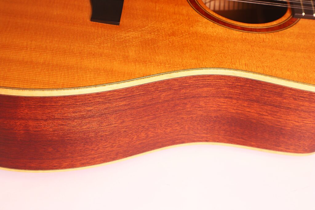 Nahaufnahme der Zarge und des Korpus der Lakewood D-14-12 Cut 2005 Akustik Gitarre, die die Holzmaserung und das helle cremefarbene Binding zeigt, wobei ein Teil der Decke und der Saiten am Rand des Bildes zu sehen ist. Nahaufnahme der Zarge und des Korpus der Lakewood D-14-12 Cut 2005 Akustik Gitarre, die die Holzmaserung und das helle cremefarbene Binding zeigt, wobei ein Teil der Decke und der Saiten am Rand des Bildes zu sehen ist.