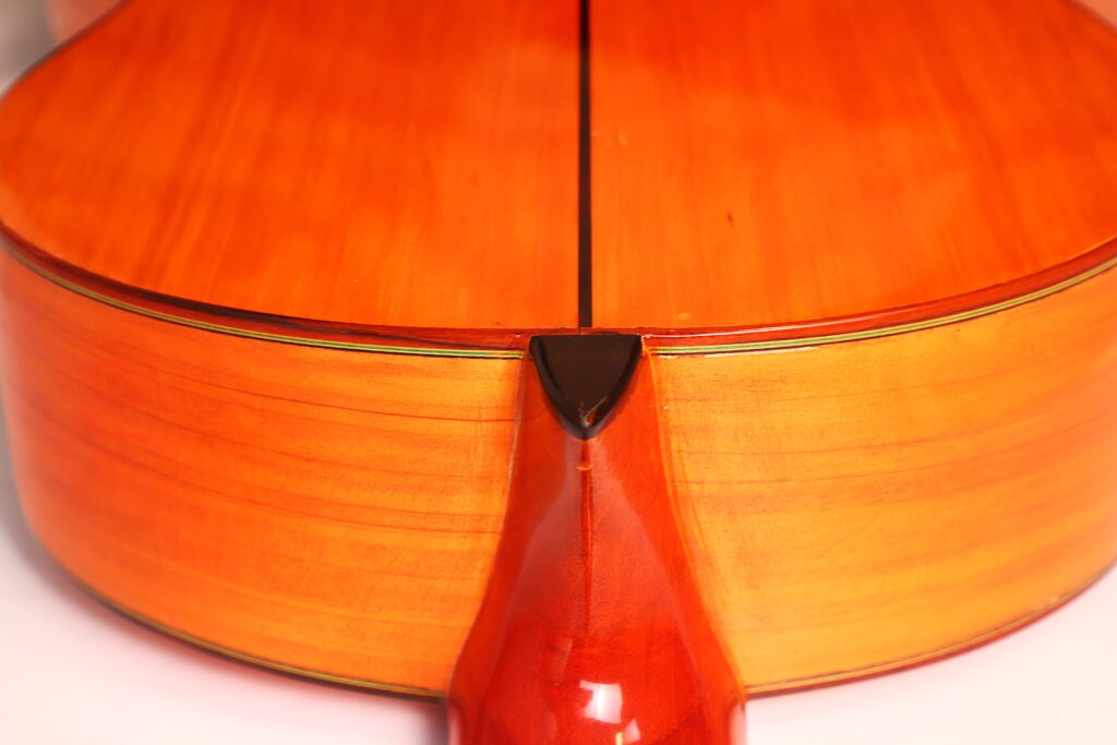 Nahaufnahme der Rückseite und der Seite eines orange-braunen Saiteninstruments mit sichtbarer Holzmaserung und glänzender Lackierung – ein Spiegelbild der handwerklichen Kunstfertigkeit, die auch in der José M Palmero 2023 Flamenco-Gitarre zum Ausdruck kommt. Nahaufnahme der Rückseite und der Seite eines orange-braunen Saiteninstruments mit sichtbarer Holzmaserung und glänzender Lackierung – ein Spiegelbild der handwerklichen Kunstfertigkeit, die auch in der José M Palmero 2023 Flamenco-Gitarre zum Ausdruck kommt.