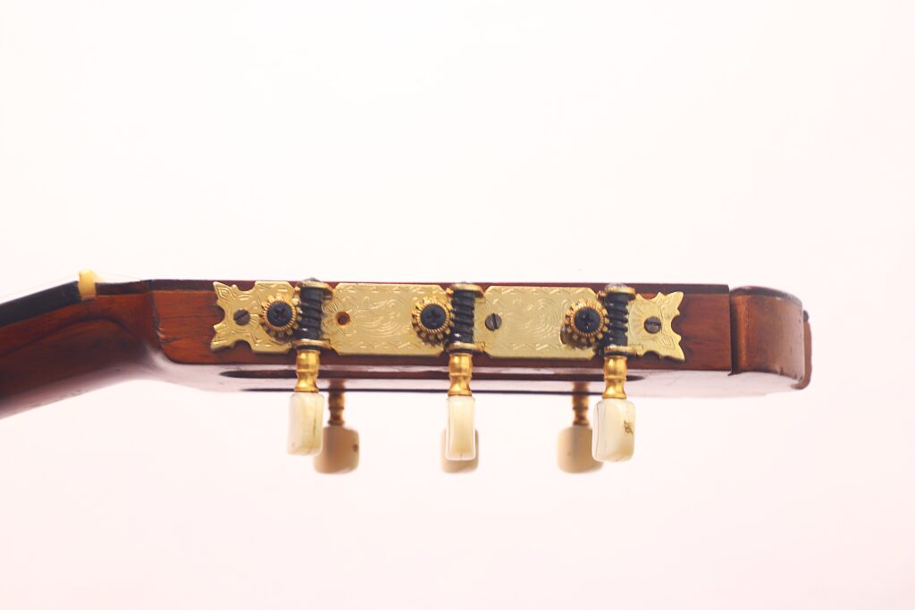 Seitliche Nahaufnahme der Stimmwirbel und der Kopfplatte einer Flamenco-Gitarre von Jose Ramirez aus dem Jahr 1955 mit drei goldfarbenen Stimmknöpfen und elfenbeinfarbenen Knöpfen vor einem schlichten hellen Hintergrund. Seitliche Nahaufnahme der Stimmwirbel und der Kopfplatte einer Flamenco-Gitarre von Jose Ramirez aus dem Jahr 1955 mit drei goldfarbenen Stimmknöpfen und elfenbeinfarbenen Knöpfen vor einem schlichten hellen Hintergrund.