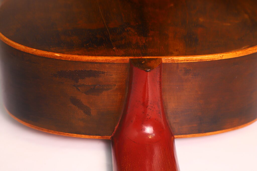 Nahaufnahme des Bodens und der Halsverbindung einer Andres Marin 1900 Klassische Gitarre, die den glänzenden roten Hals und den abgenutzten, dunkelbraunen Holzkorpus mit sichtbarer Maserung und Gebrauchsspuren zeigt. Nahaufnahme des Bodens und der Halsverbindung einer Andres Marin 1900 Klassische Gitarre, die den glänzenden roten Hals und den abgenutzten, dunkelbraunen Holzkorpus mit sichtbarer Maserung und Gebrauchsspuren zeigt.