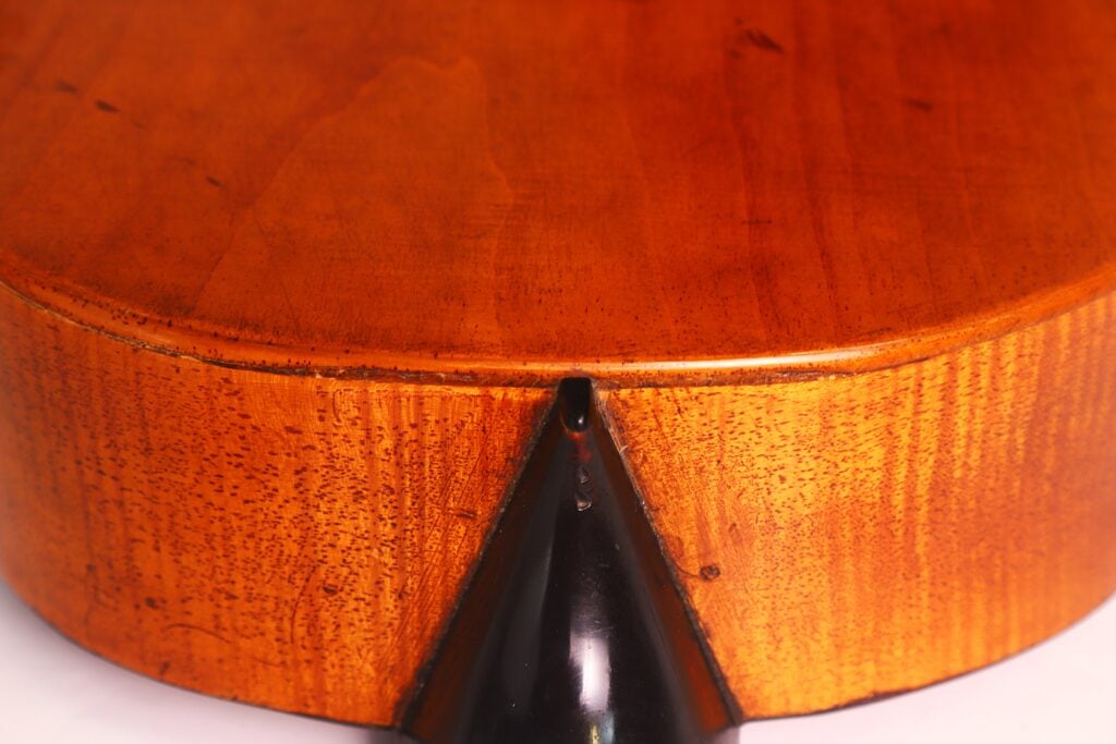 Nahaufnahme der Unterkante einer Romantikgitarre von Stephan Thumhart aus dem Jahr 1838, die die glatte, lackierte Holzmaserung und den mittig angebrachten schwarzen Saitenhalter hervorhebt. Nahaufnahme der Unterkante einer Romantikgitarre von Stephan Thumhart aus dem Jahr 1838, die die glatte, lackierte Holzmaserung und den mittig angebrachten schwarzen Saitenhalter hervorhebt.