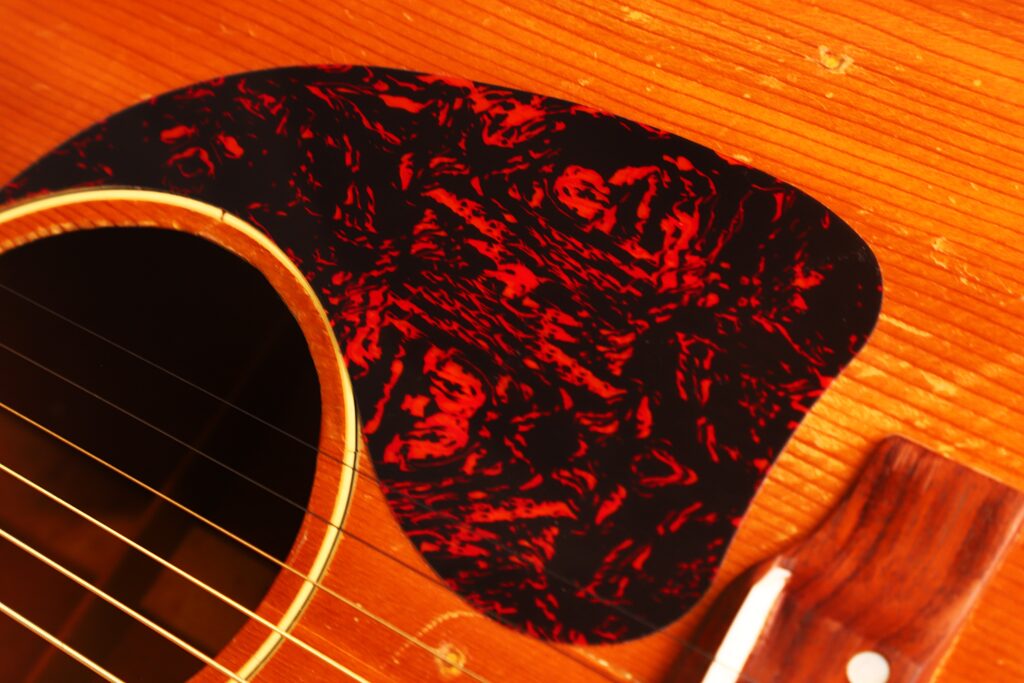 Nahaufnahme der Gibson J-50 Akustikgitarre aus dem Jahr 1952, die ihren Holzkorpus, das Schallloch, die Saiten und das markante, marmorierte rot-schwarze Schlagbrett zeigt. Nahaufnahme der Gibson J-50 Akustikgitarre aus dem Jahr 1952, die ihren Holzkorpus, das Schallloch, die Saiten und das markante, marmorierte rot-schwarze Schlagbrett zeigt.