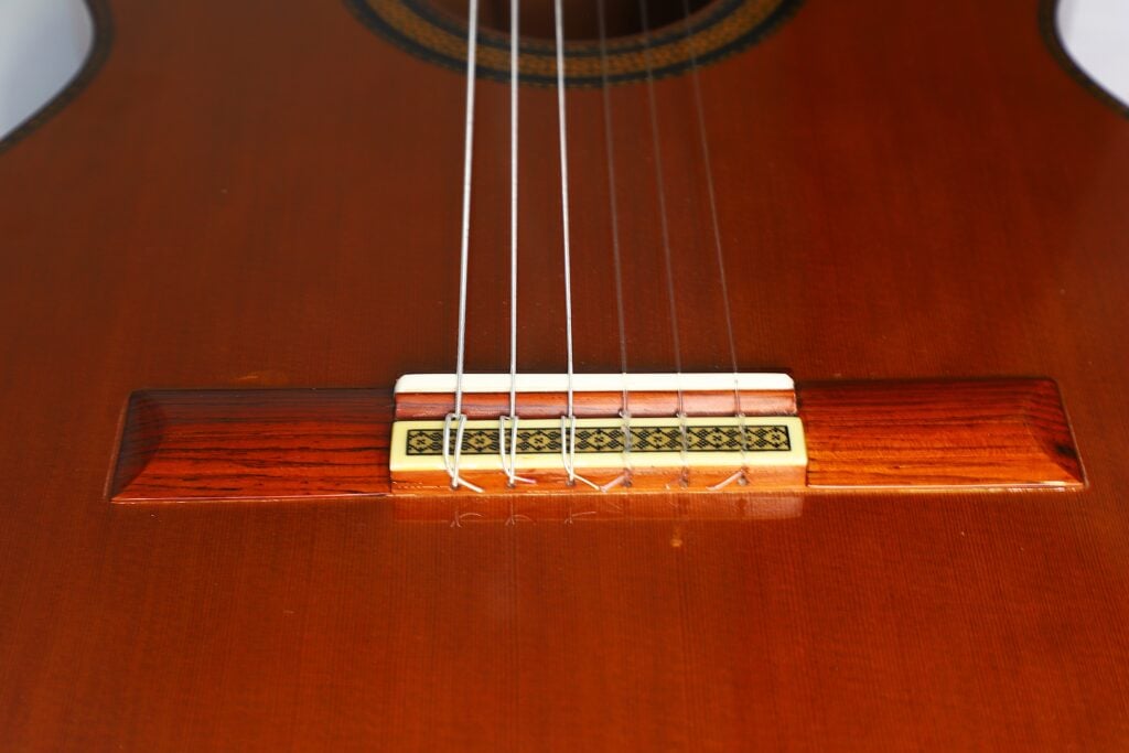 Nahaufnahme des Saitenhalters der „Doble Tapa Klassische Gitarre“ von Manuel Contreras aus dem Jahr 1981, bei der sechs Saiten über einem Holzsteg verlaufen, der mit dekorativen Einlagen verziert ist und auf dem polierten braunen Korpus sitzt. Nahaufnahme des Saitenhalters der „Doble Tapa Klassische Gitarre“ von Manuel Contreras aus dem Jahr 1981, bei der sechs Saiten über einem Holzsteg verlaufen, der mit dekorativen Einlagen verziert ist und auf dem polierten braunen Korpus sitzt.