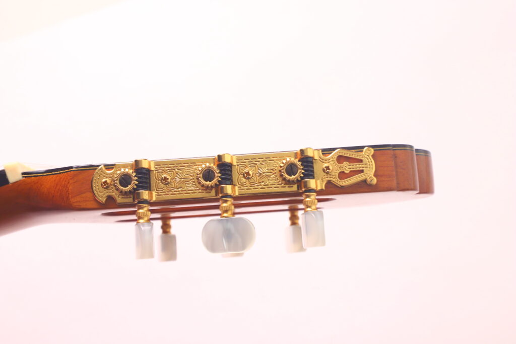 Seitliche Nahaufnahme der Kopfplatte der Vicente Carrillo 1998 Torres Klassische Gitarre, die verschnörkelte goldene Stimmwirbel mit weißen Knöpfen vor einem rosa Hintergrund zeigt. Seitliche Nahaufnahme der Kopfplatte der Vicente Carrillo 1998 Torres Klassische Gitarre, die verschnörkelte goldene Stimmwirbel mit weißen Knöpfen vor einem rosa Hintergrund zeigt.