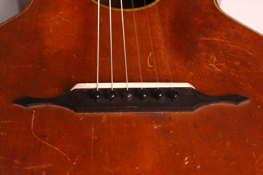 Nahaufnahme des Stegs und der Saiten der Braun & Hauser 1905 Klassische Gitarre, die eine abgenutzte braune Holzoberfläche mit sichtbaren Kratzern und einen dekorativen schwarzen Steg mit sechs Saiten aufweist. Nahaufnahme des Stegs und der Saiten der Braun & Hauser 1905 Klassische Gitarre, die eine abgenutzte braune Holzoberfläche mit sichtbaren Kratzern und einen dekorativen schwarzen Steg mit sechs Saiten aufweist.