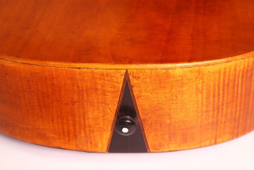 Nahaufnahme der Unterkante einer „Romantikgitarre“ von Stephan Thumhart aus dem Jahr 1838, mit glänzender Lackierung, sichtbarer Maserung, einer dreieckigen schwarzen Einlage und einem Gurtknopf mit weißem Punkt. Nahaufnahme der Unterkante einer „Romantikgitarre“ von Stephan Thumhart aus dem Jahr 1838, mit glänzender Lackierung, sichtbarer Maserung, einer dreieckigen schwarzen Einlage und einem Gurtknopf mit weißem Punkt.