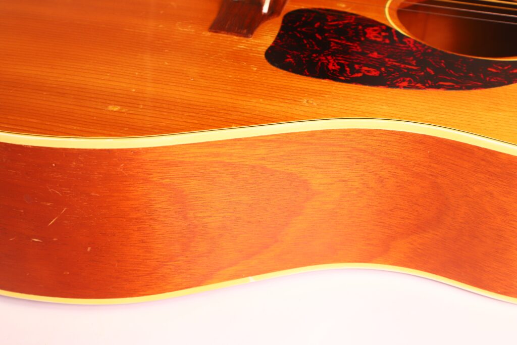 Nahaufnahme des Korpus der Gibson J-50 Akustikgitarre aus dem Jahr 1952, bei der die Holzmaserung, die cremefarbene Einfassung und das Schlagbrett besonders gut zur Geltung kommen. Oben rechts sind die Saiten und das Schallloch teilweise zu sehen. Nahaufnahme des Korpus der Gibson J-50 Akustikgitarre aus dem Jahr 1952, bei der die Holzmaserung, die cremefarbene Einfassung und das Schlagbrett besonders gut zur Geltung kommen. Oben rechts sind die Saiten und das Schallloch teilweise zu sehen.